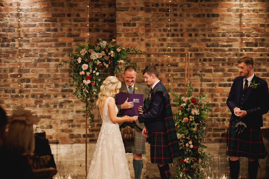 Bride puts grooms ring on his finger whilst celebrant looks on smiling